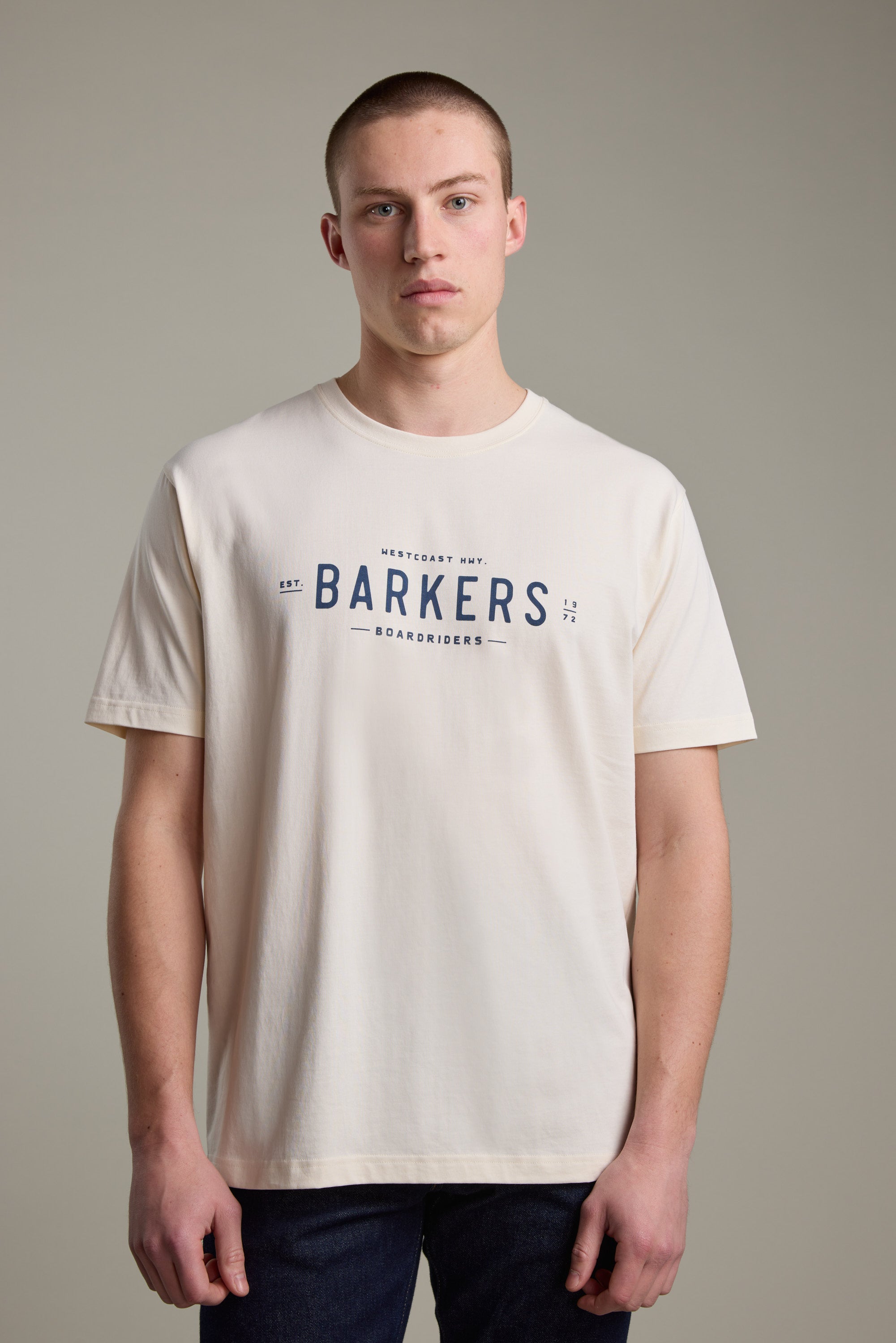 A young man with short hair wears the Barkers Boardriders Logo Tee, a light-colored organic cotton shirt featuring the brand’s logo on the front, standing against a neutral background.
