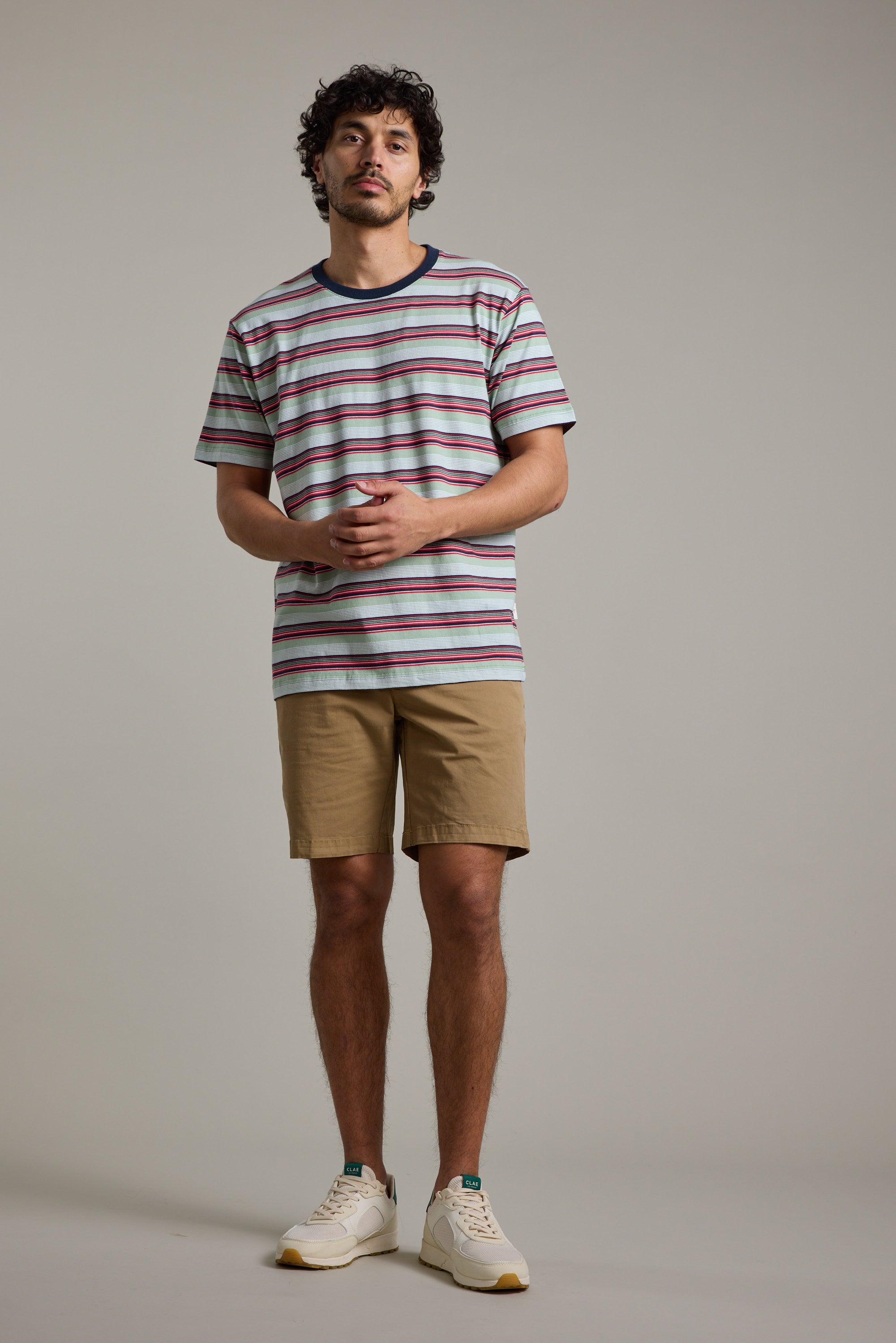 A man with curly hair and a slight beard stands against a plain backdrop, wearing the Barkers Alvin Stripe Tee with a crew neck, tan shorts, and white sneakers, his hands loosely clasped in front of him.
