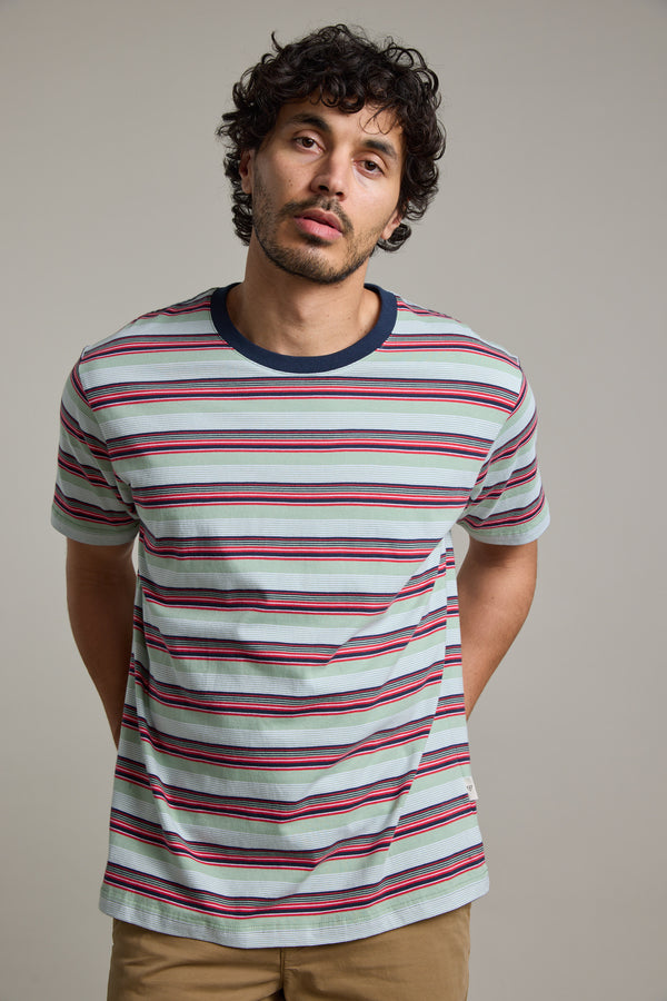 A man with curly dark hair and a short beard wears the Barkers Alvin Stripe Tee—an organic cotton crewneck with red, navy, and white stripes—paired with tan pants. He stands against a plain gray background, hands behind his back.