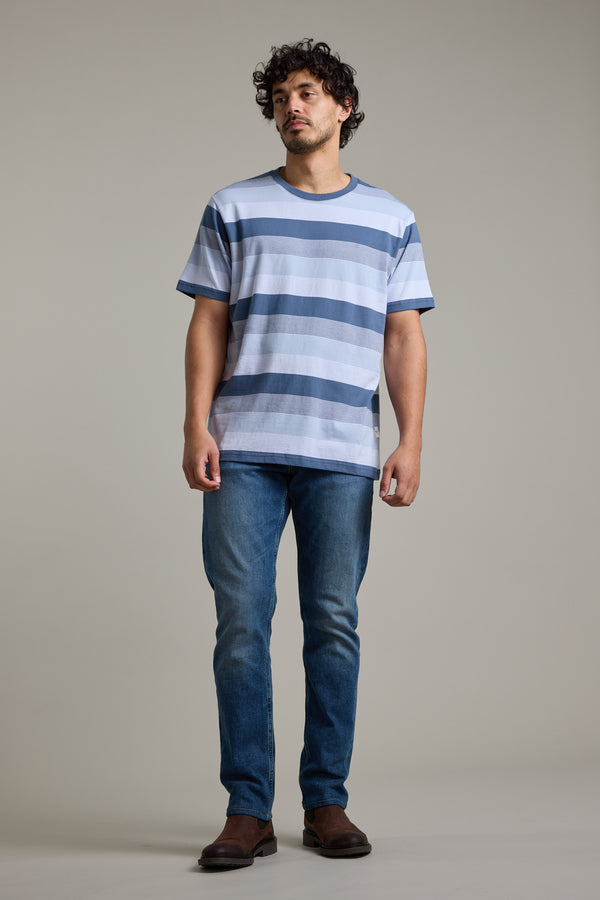 A man with curly hair wears the Barkers Fitzroy Stripe Tee, blue jeans, and brown boots, standing against a neutral gray background.
