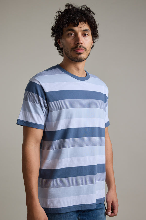 A man with curly dark hair and a short beard is wearing the Barkers Fitzroy Stripe Tee with blue jeans, standing in front of a plain, light-colored background.