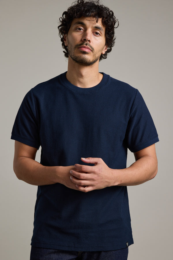 A man with curly dark hair and a mustache wears the Barkers Signature Knit Tee in dark blue, featuring ribbed cuffs. He stands against a plain light background, looking at the camera with his hands clasped in front of him.
