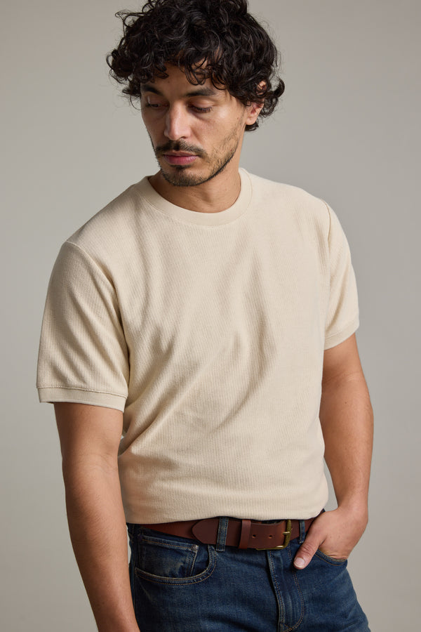 A man with curly dark hair and facial hair wears the Barkers Signature Knit Tee in beige, paired with blue jeans and a brown belt. He looks downward with one hand in his pocket against a plain background.