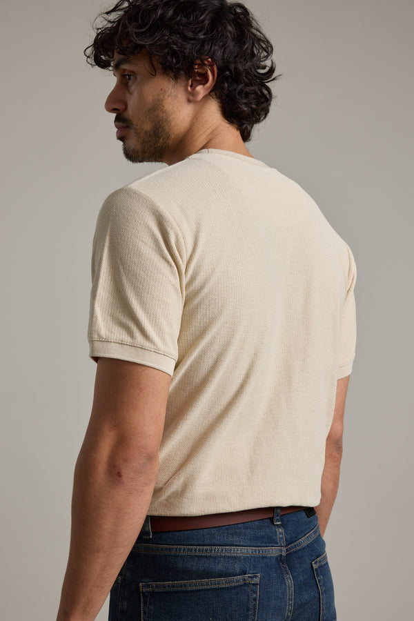 A man with curly dark hair models the Barkers Signature Knit Tee, a light beige short-sleeve shirt, paired with blue jeans. He stands turned slightly away from the camera against a plain gray background.
