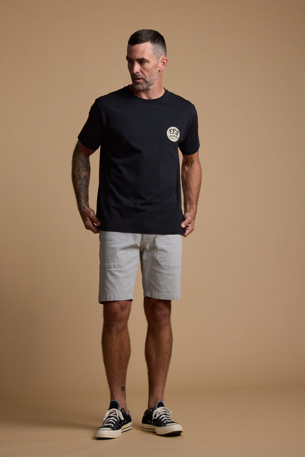 A man with short hair and tattoos wears the Legasea Tail Print Tee by Legasea, paired with light gray shorts and black low-top sneakers. He stands against a plain beige background, looking slightly to his left.