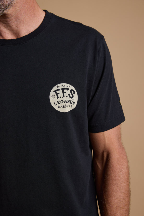 A man in a black Legasea Tail Print Tee, featuring a round white “F.F.S LEGASEA BARKERS” logo and inspired by the New Zealand coastline, stands against a plain beige background with only part of his face and upper body visible.