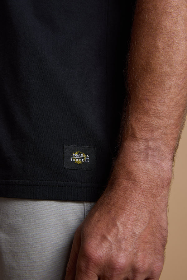 A close-up of a person's right arm and hand in light-colored pants features the Legasea Tail Print Tee by Legasea, a black organic cotton shirt with a small branded label near the hem.