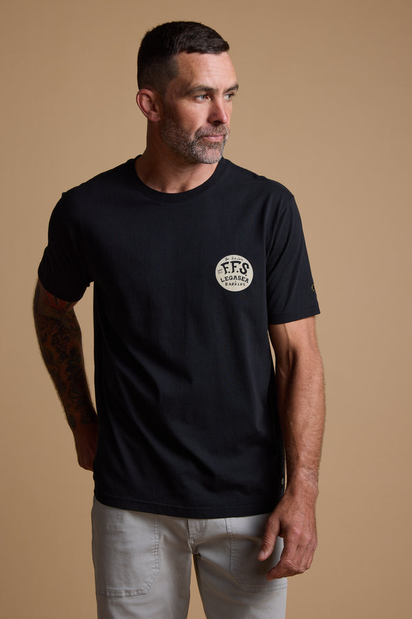 A man with short dark hair and a beard wears the Legasea Tail Print Tee—a black organic cotton T-shirt by Legasea with a small circular logo on the chest—and light-colored pants, standing against a plain beige background.