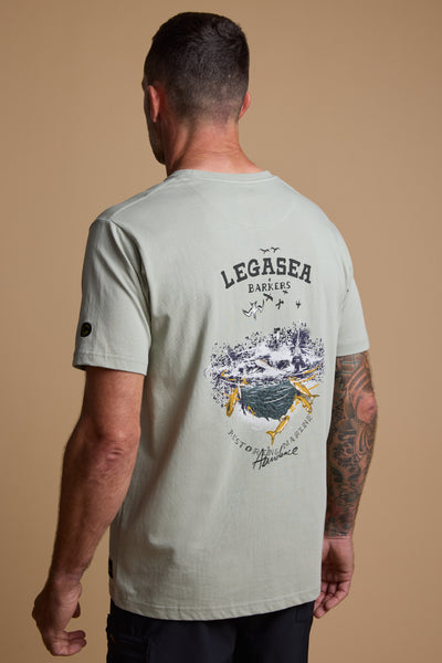A man with short hair and a tattooed arm wears the Legasea Workup Print Tee by Legasea, a light gray organic cotton shirt featuring a large back graphic with "LEGASEA BARKERS" and an illustrated ocean scene with birds and fish.