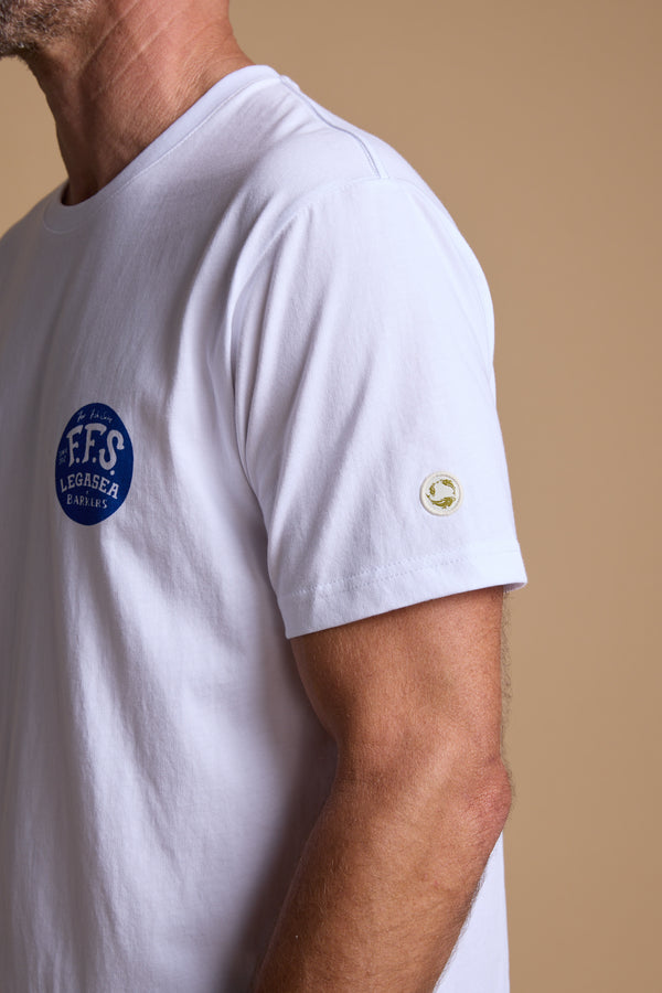 A man wears the Legasea Marlin Print tee—white organic cotton, blue circular chest logo, and sleeve patch—standing against a beige background. Legasea design inspired by the New Zealand coastline blends comfort and style.