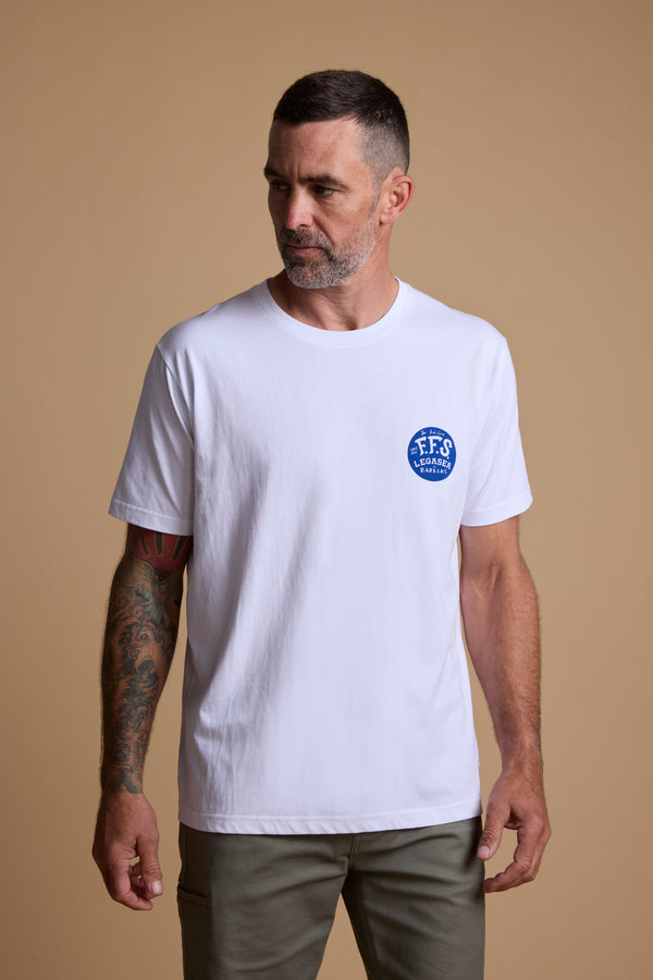 A bearded man with short hair and colorful left-arm tattoos wears the Legasea Marlin Print tee, featuring a small blue circular logo on the left chest, by Legasea. He stands against a beige background looking to the side.