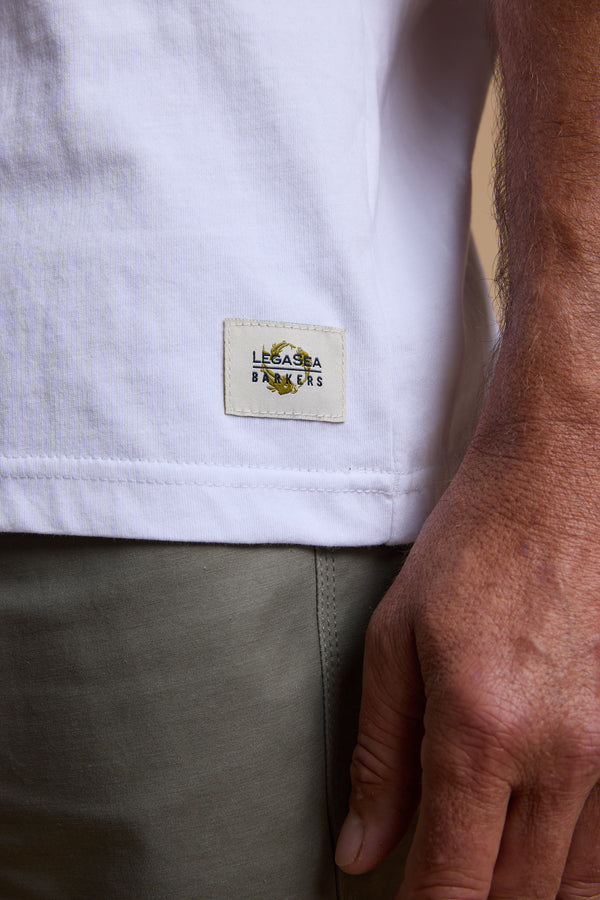 A close-up of a person wearing the Legasea Marlin Print tee, featuring organic cotton and a Legasea Bankers label by the hem, paired with olive green pants. Their relaxed pose captures the laid-back vibe of the New Zealand coastline.