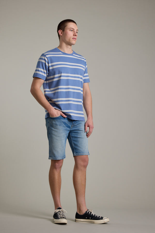 A young man stands against a plain background, wearing the Barkers Northway Stripe Tee—blue and white organic cotton stripes—perfect for summer. He pairs it with light blue denim shorts and black sneakers, one hand in his pocket as he looks off to the side.