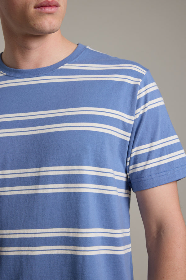 A person wears the Barkers Northway Stripe Tee, featuring blue and white horizontal stripes, shown from shoulders to above the elbow against a plain background with a partially visible face—a timeless summer essential.
