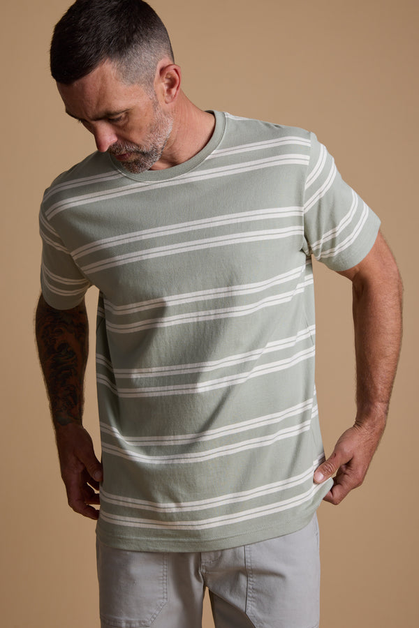 A man with short dark hair and a beard wears the Barkers Northway Stripe Tee and light gray pants, looking down as he adjusts his shirt against a beige background—the perfect summer wardrobe choice.