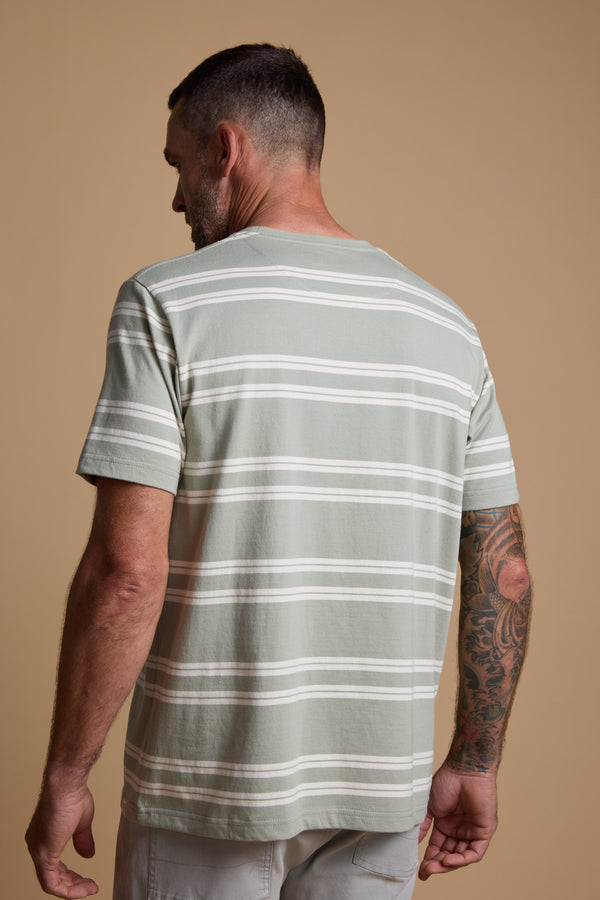 A man with short dark hair and a tattooed right arm is seen from the back, wearing the Barkers Northway Stripe Tee—an organic cotton light green striped shirt—against a plain beige background, perfect for summer.