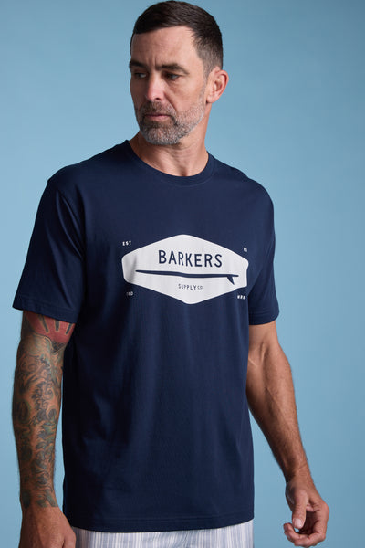 A man with short dark hair and a trimmed beard wears a navy Barkers Supply Co Print Tee and light striped pants, standing against a light blue background. He has a tattoo sleeve on his left arm and is looking to his left.
