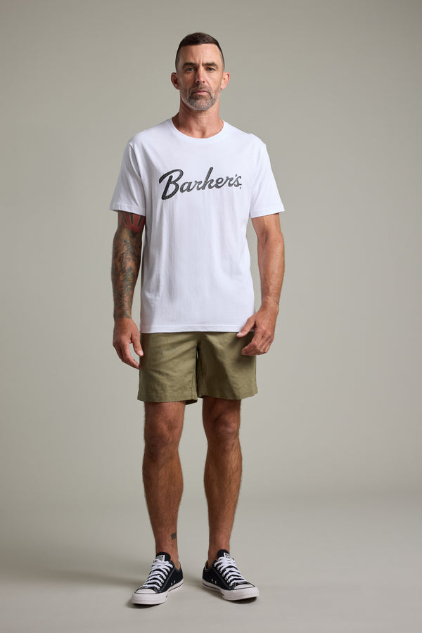 A man with short hair, a beard, and tattoos on his left arm faces forward in a plain light grey setting, wearing a white Barkers Finely Tuned Print Tee, olive green shorts, and black sneakers.