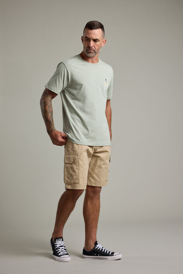 A man with short hair and tattoos wears the Barkers BMC Sound Sessions Print Tee, light green in color, paired with beige cargo shorts and black sneakers. He stands against a gray background, looking left with one hand in his pocket.