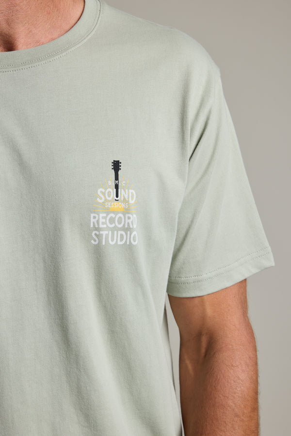 A person wears the Barkers BMC Sound Sessions Print Tee—a light gray organic cotton shirt with a small guitar graphic and "SOUND SESSIONS RECORD STUDIO" text on the left chest, giving vintage band merch vibes. Only the upper torso is visible.