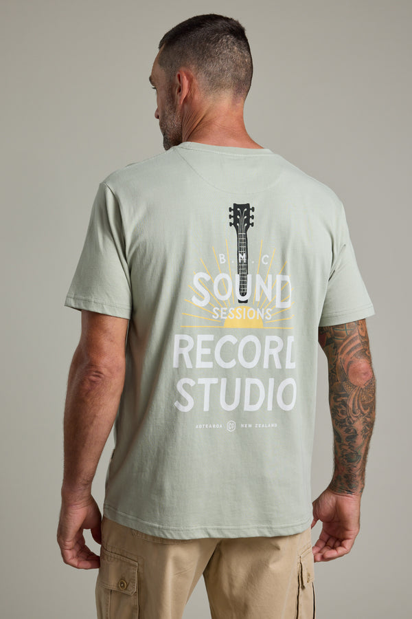 A man with short hair and tattoos on his right arm wears the Barkers BMC Sound Sessions Print Tee—a regular fit, light sage organic cotton shirt featuring a guitar headstock and “SOUND SESSIONS RECORD STUDIO” graphic on the back.