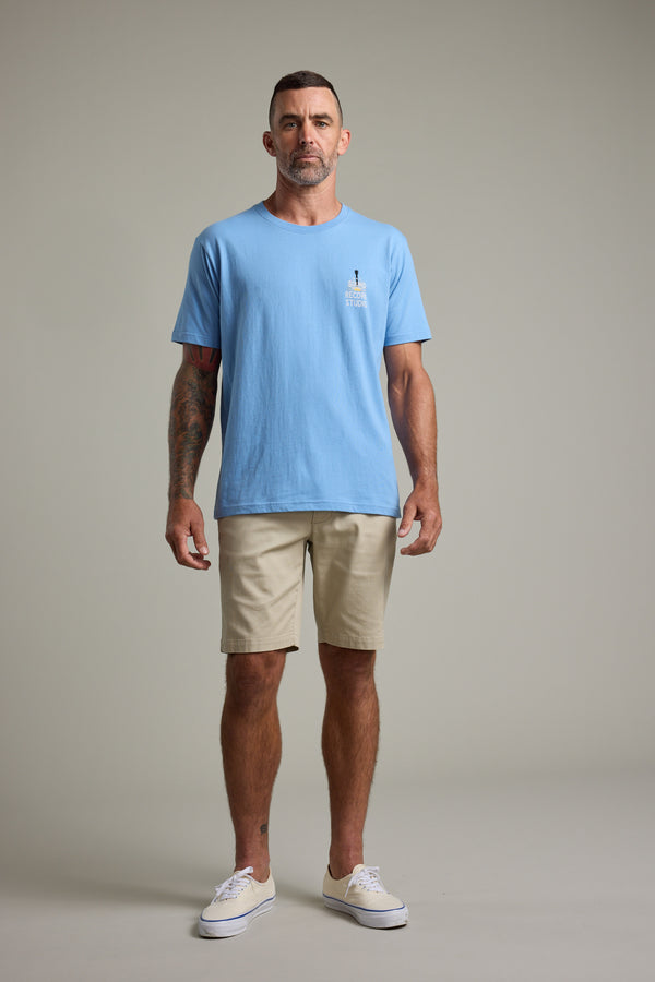 A man with short hair and visible arm tattoos stands facing forward against a plain background, wearing the Barkers BMC Sound Sessions Print Tee in light blue, beige shorts, and white sneakers. He has a neutral expression.