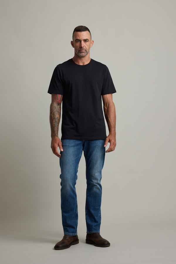 Man in Barkers Good Earth Cotton Essential Tee and blue jeans stands against a plain background.