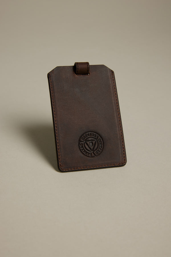 The Barkers Voyage Leather Luggage Tag stands upright against a plain beige background—crafted in brown leather with stitched edges and an embossed circular logo, it’s the perfect corporate gift or stylish travel accessory.