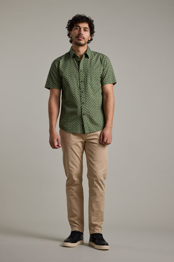 A man stands against a plain background wearing the Barkers Oxnard Print Shirt in green organic cotton, paired with beige pants and black sneakers. He looks forward with a neutral expression and relaxed posture.