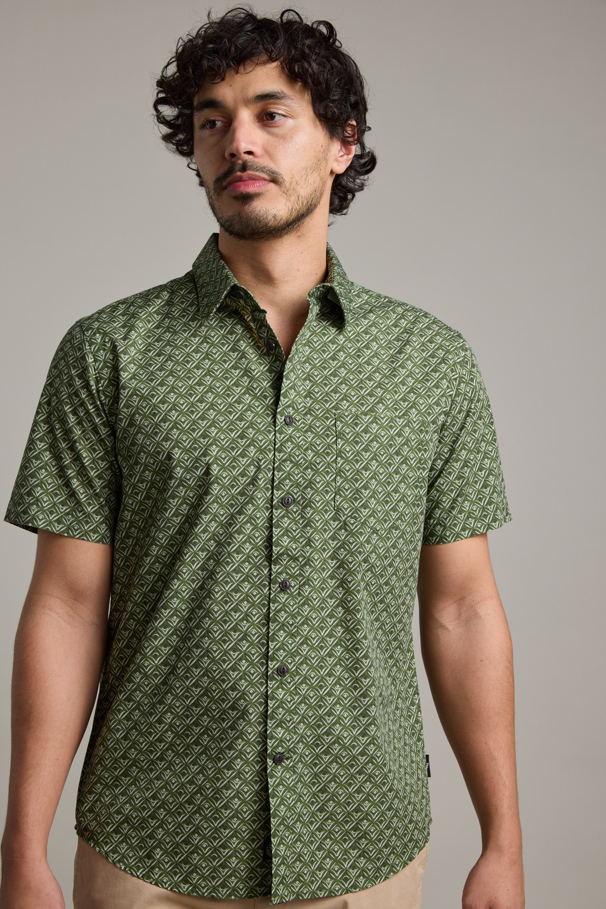 A man with short curly hair and facial hair models the Barkers Oxnard Print Shirt, a green, short-sleeve shirt made from organic cotton, standing against a plain light gray background.