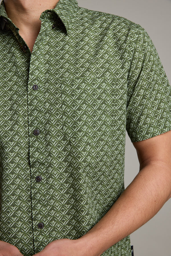 A person is shown from the torso up wearing the Barkers Oxnard Print Shirt, a green short sleeve shirt with a white geometric pattern, made from organic cotton, set against a plain background.