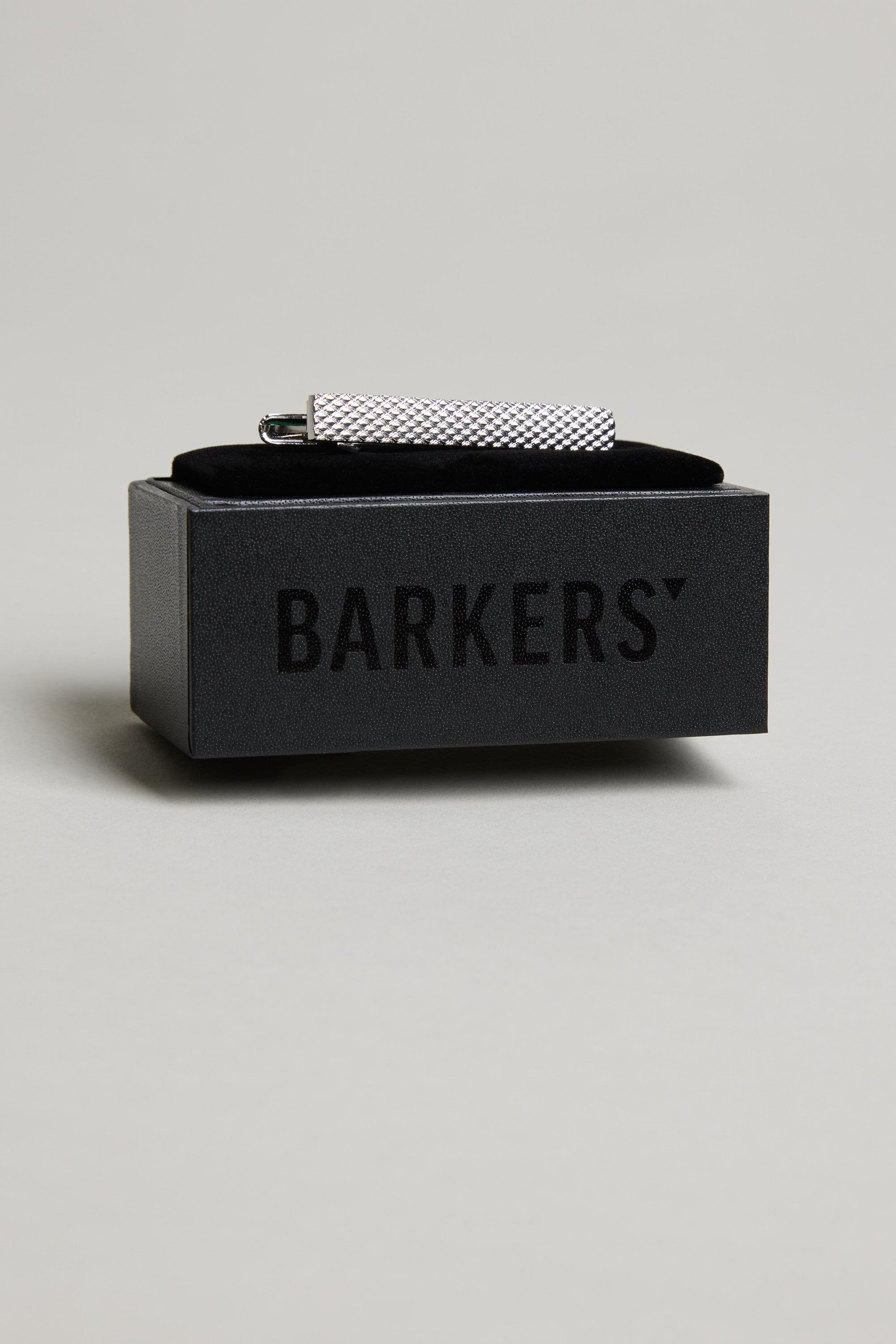 The Barkers Texture Tie Clip, featuring a textured silver-tone design, is displayed on a black box with the brand's logo, standing out against a light grey background.