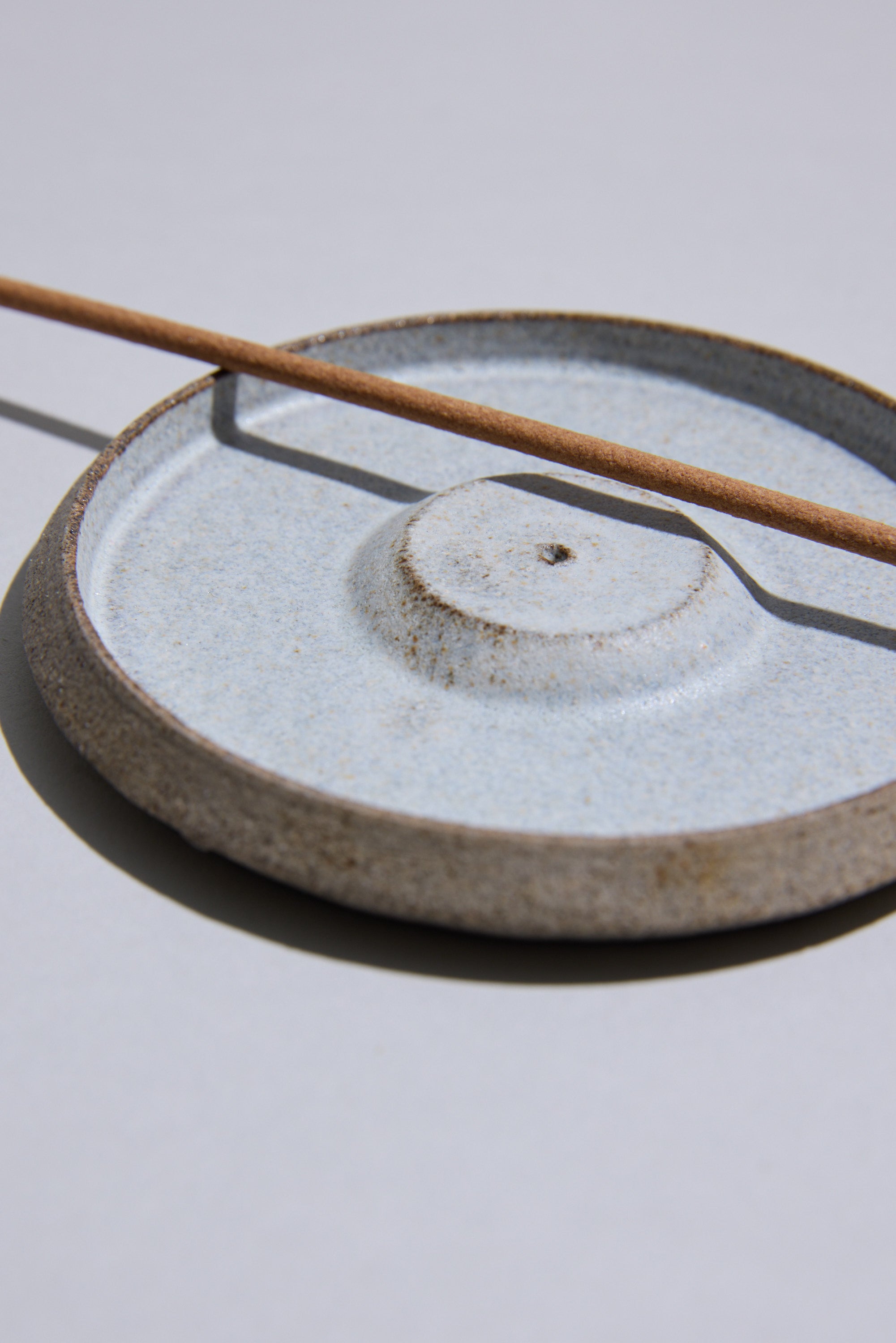 The Studio Foon Incense Dish by Other is a light-colored, handcrafted ceramic piece from New Zealand with a round base, shown holding a single stick of brown incense on a plain, light background.