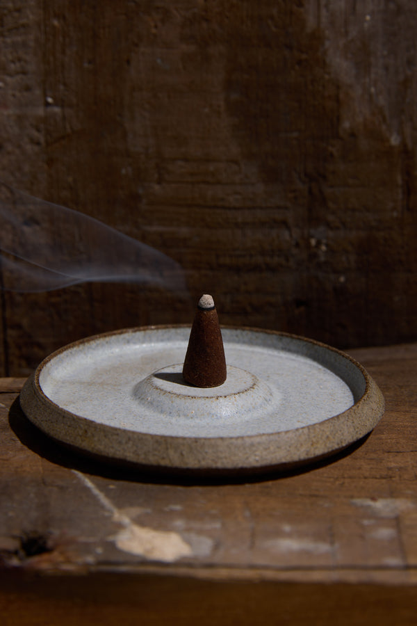 A single incense cone burns on the handcrafted Studio Foon Incense Dish by Other, releasing a delicate wisp of smoke against a rustic wooden background.