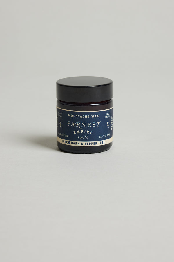 A small black jar of Earnest Empire Moustache Wax by Other, with a blue label reading Birch Bark & Pepper Tree and made from natural ingredients, sits on a plain light gray background.