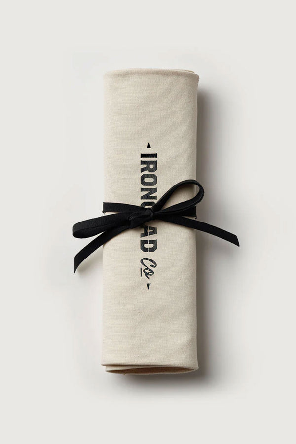The IronClad Ghost Knife Roll: a cream-colored canvas napkin, rolled and tied with a black ribbon, featuring the IronClad logo in black on the front. Displayed against a plain, light gray background.