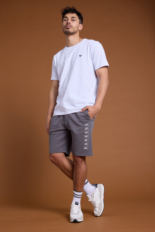 A man stands against a plain brown background, wearing a plain white t-shirt and Barkers Organic Classic Print Track Shorts in gray. He pairs them with white socks and sneakers, hands in his pockets, looking slightly upward.