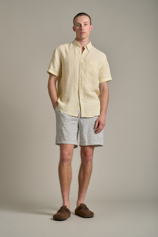 A man stands against a plain background wearing the Barkers Gentry Linen Shirt in light yellow, light gray striped shorts, and brown loafers, hands in pockets—a breathable choice for any summer wardrobe.