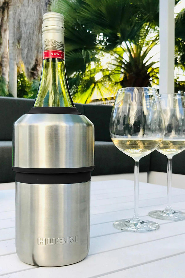 A Huski Wine Cooler by Huski chills a wine bottle on a white outdoor table with two filled glasses, set against green palm trees and a patio—keeping your wine cold for hours and perfect for relaxing outside.