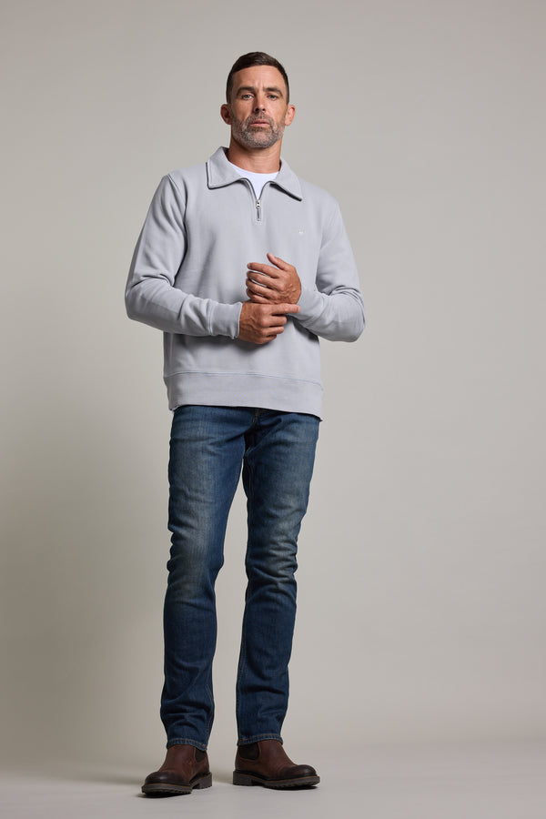 A man stands before a plain light background, wearing a Barkers Galway GD 1/4 Zip in light gray, paired with blue jeans and brown shoes. He gazes ahead with a neutral expression, hands clasped loosely in front of him.