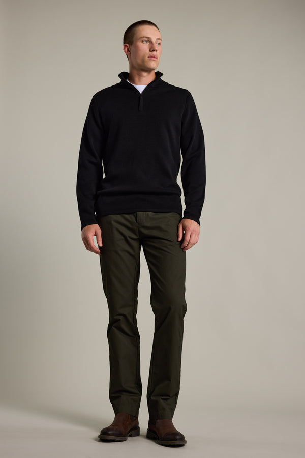 A man faces slightly to the side, wearing a black Barkers Brandon ZQRX Merino 1/4 Zip pullover, olive green pants, and brown shoes against a plain light background—ideal for cold-weather layering.