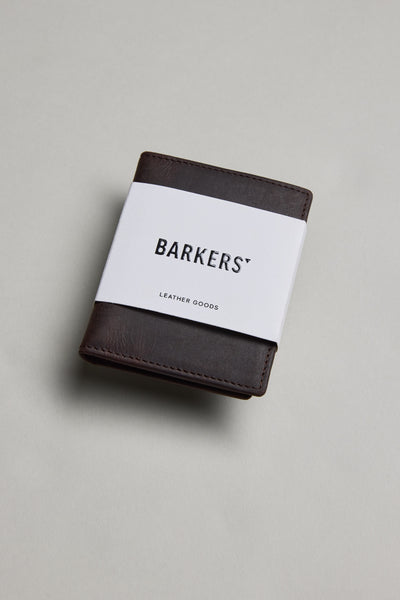 A Marlin Leather Slim Wallet by Barkers, dark brown and crafted from premium cowhide, is wrapped in a white paper band labeled "Barkers" and displayed on a plain light gray surface.