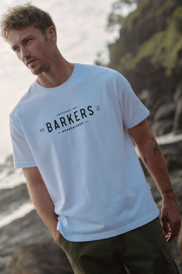 A man with short, light brown hair and stubble stands by a rocky coast, wearing the Barkers Boardriders Logo Tee—an organic cotton t-shirt—and dark olive-green shorts.