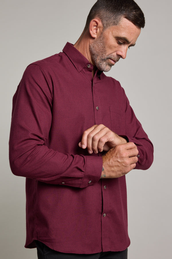 A man with short dark hair and a beard wears the Barkers Antonio Shirt, a classic fit, long-sleeved maroon style in a cotton-wool blend. He looks down while fastening his cuff against a neutral background.