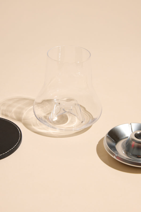 The Other Whisky Glass Individual, with a unique shape, rests on a beige surface beside a round black coaster and silver metal dish, all casting soft shadows.