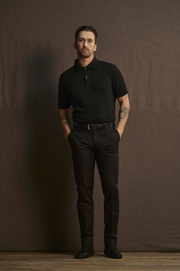 A man with short dark hair and forearm tattoos stands against a brown backdrop, hands in pockets, wearing Barkers Lyndon Cotton Pants with a black polo shirt and black shoes for a sharp smart casual look.