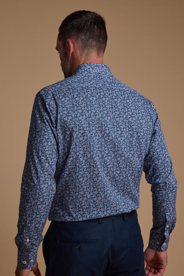 A man stands with his back to the camera, wearing the Barkers Watkins Print Business Shirt—tailored fit, long-sleeved, and blue patterned—tucked into dark blue pants, set against a plain beige background.