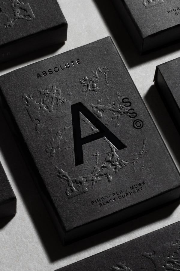 A close-up of Solid State - Absolute, a pocket-sized black box cologne embossed with an "A," textured botanical motifs, and the notes "Pineapple, Musk, Black Currant." Creed Aventus-inspired scent by Solid State; other boxes surround it.