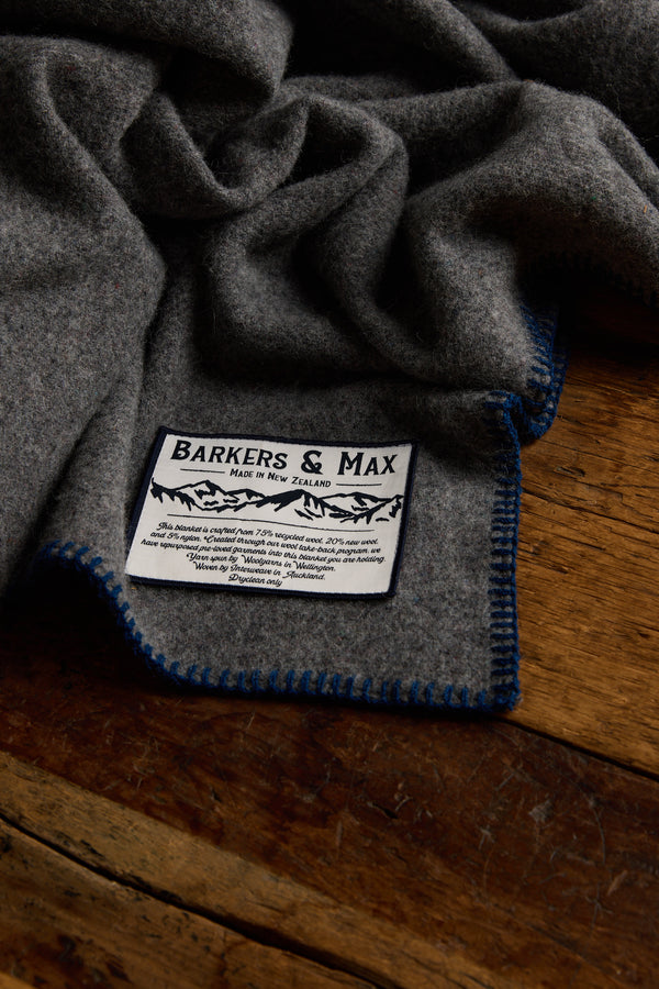 A grey Wool Take Back Blanket by Other, made from sustainable wool with blue edge stitching and a white label, is shown lying on a wooden surface.