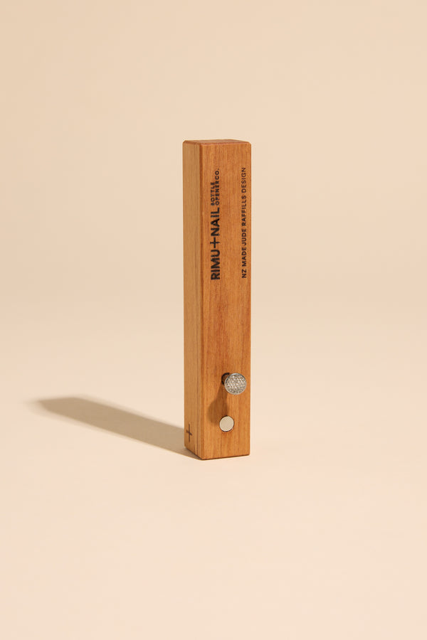 The Rimu & Nail Bottle Opener by Other stands upright on a beige surface, featuring a round metal knob, a small button, and engraved details on the front—showcasing its unique, handmade NZ design.